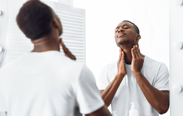 Unrasierte schwarze Mann berühren Hals in der Nähe von Spiegel im Badezimmer - Foto, Bild