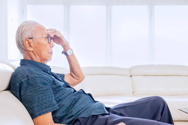 Side view of senior man suffering headache while sitting on the couch in the living room. Shot at home - 写真・画像