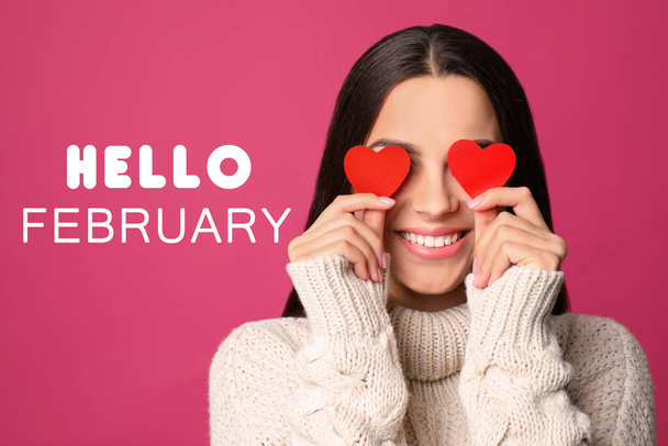 Greeting card with text Hello February. Young woman holding paper hearts near eyes on pink background - Photo, Image