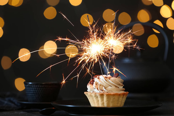 Cupcake with burning sparkler on black table against blurred festive lights - 写真・画像