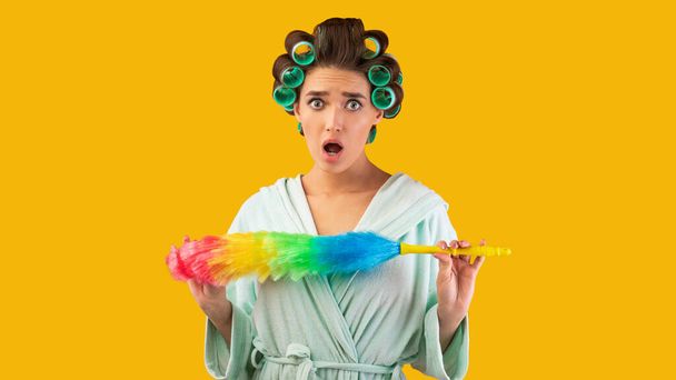 Shocked Housewife Holding Feather Duster Standing Over Yellow Studio Background - Foto, Imagen