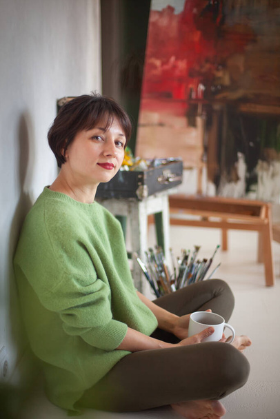 Young caucasian artist woman having rest and drinking hot tea near painted oil painting on easel in her workshop. Selective focus. Art and craft. Hobby and leisure. Creative people. - Foto, imagen