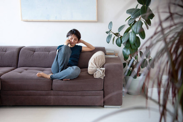 Young woman with short hair style siting on couch and talking on phone at home. Housewife relaxing and chatting through mobile phone. Interior design. Copy space. - Fotó, kép