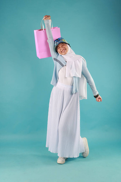Portrait of an excited beautiful girl wearing dress holding shopping bags isolated over blue background. - Photo, Image