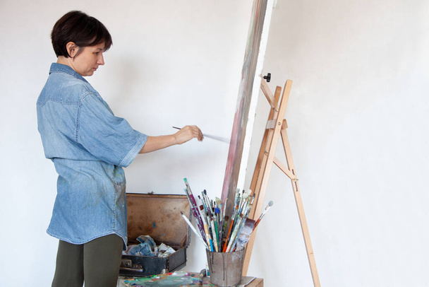 Young woman artist holding paintbrush and painting with oil paint on framed cotton canvas on easel at home. Hobby and leisure concept. Painter creating art. Interior. Copy space. - Fotoğraf, Görsel