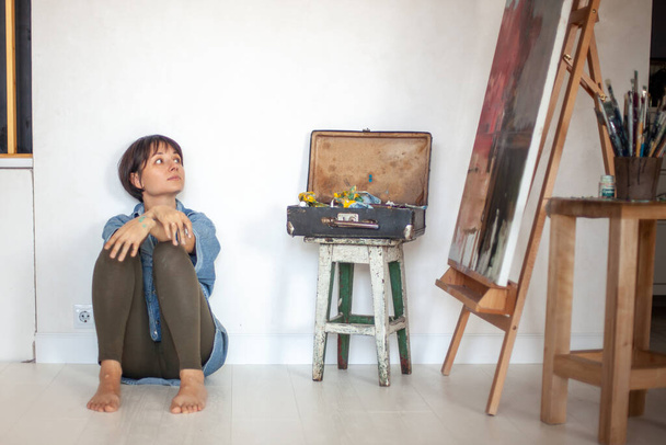 Female artist with paintbrush sitting, looking to painted oil painting on easel near vintage wooden box with drawing tools and bucket with paintbrushes. Painter is analysing her art creation in studio - Fotó, kép