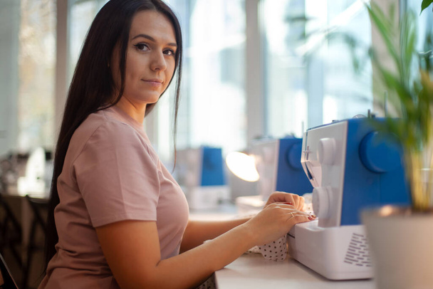 Beautiful woman tailor with long dark hair completing work with sewing machine at workplace. Neatly job. Attending lessons of seamstress. Technology in apparel industry. Manufacturing concept - 写真・画像