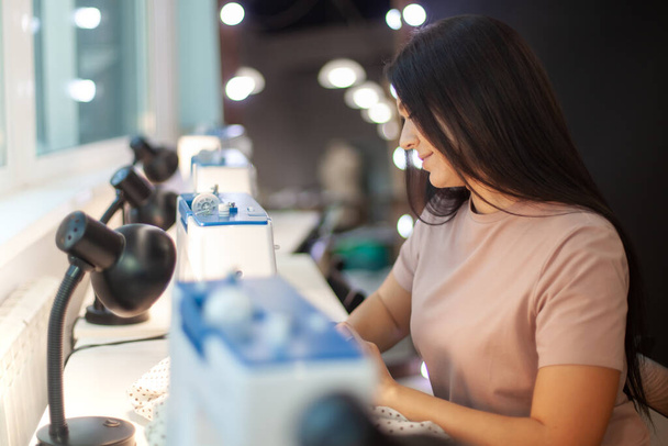 Beautiful woman tailor with long dark hair completing work with sewing machine at workplace. Neatly job. Attending lessons of seamstress. Technology in apparel industry. Manufacturing concept - 写真・画像