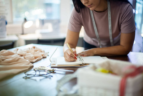 Beautiful young woman designer is making notes of clothing measurements on notebook in sewing studio. Female tailor measuring and writing work plan on notepad on desk with textile. - 写真・画像