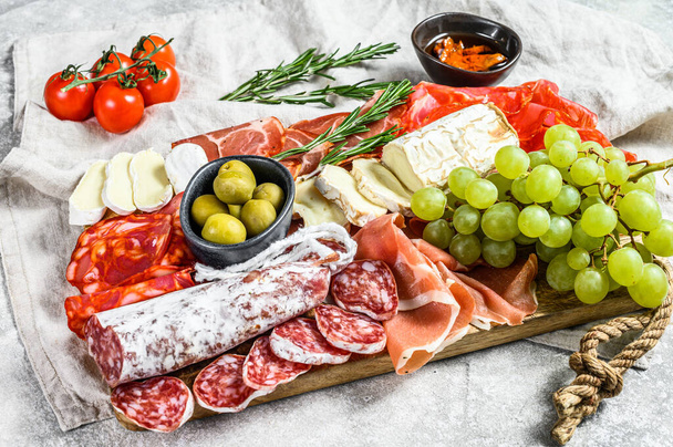 イタリアのアンティパスト、プロシクト、ハム、パルマ、ヤギ、カンベルトチーズ、オリーブ、ブドウと木製のまな板。アンティパスティ。灰色の背景。トップ表示. - 写真・画像
