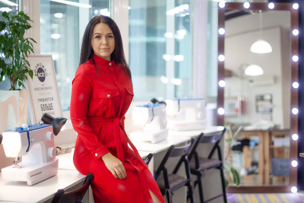 Pretty woman designer in red dress sitting on table with sewing machines in modern workshop. Elegant female entrepreneur in fashion industry. Tailor sewed and put on red dress for Christmas eve party - Valokuva, kuva