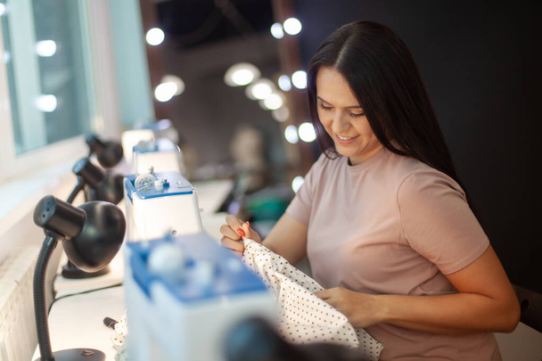 Beautiful woman tailor with long dark hair completing work with sewing machine at workplace. Neatly job. Attending lessons of seamstress. Technology in apparel industry. Manufacturing concept - Fotoğraf, Görsel