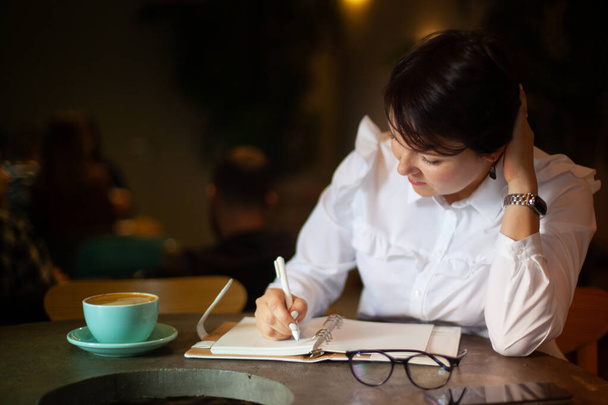woman in business white blouser  of coffee shop and writing a Christmas shopping list with cup of blue latte. Planning Christmas holidays. Organising and planning concept. - 写真・画像