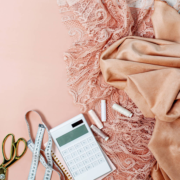Top view of pink coral coloured lace and silk fabrics with thread spools, calculator on notepad, tape measure, tailoring scissors near cup of coffee on pink background. Evening gowns sewing concept. - Zdjęcie, obraz