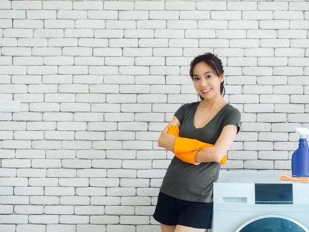 Mooie glimlachende Aziatische vrouw, gelukkig huisvrouw dragen oranje beschermende rubberen handschoenen staan met armen gevouwen naast wasmachine op witte baksteen muur achtergrond in wasruimte met kopieerruimte. - Foto, afbeelding