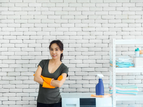 Mooie glimlachende Aziatische vrouw, gelukkig huisvrouw dragen oranje beschermende rubberen handschoenen staan met armen gevouwen naast wasmachine op witte baksteen muur achtergrond in heldere schone wasruimte. - Foto, afbeelding