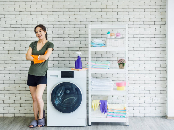 Beautiful smiling Asian woman, happy housewife wearing orange protective rubber gloves standing with arms folded beside washing machine on white brick wall background in bright clean laundry room. - Photo, Image