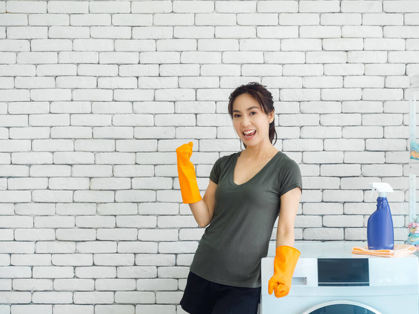 Happy uśmiechnięta Azjatka, gospodyni domowa nosząca pomarańczowe gumowe rękawiczki podnosząca rękę w zwycięskim geście i świętująca sukces w pobliżu pralki na ceglanym tle ściany w pralni z przestrzenią do kopiowania. - Zdjęcie, obraz