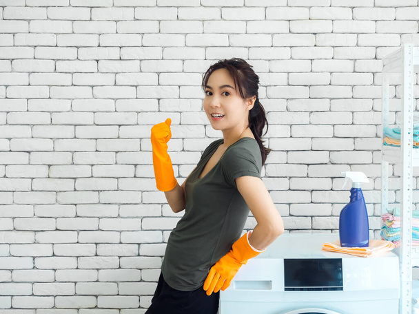 Onnellinen hymyilevä aasialainen nainen, kotiäiti yllään oranssi kumi käsineet seisoo ja nostaa kätensä voittaa ele ja juhlii menestystä lähellä pesukone tiiliseinä tausta pesutupa. - Valokuva, kuva