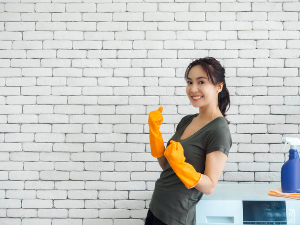Feliz mujer asiática sonriente, ama de casa con guantes de goma levantando puños en el gesto ganador y celebrando el éxito cerca de la lavadora en el fondo de la pared de ladrillo en la sala de lavandería con espacio para copias. - Foto, imagen