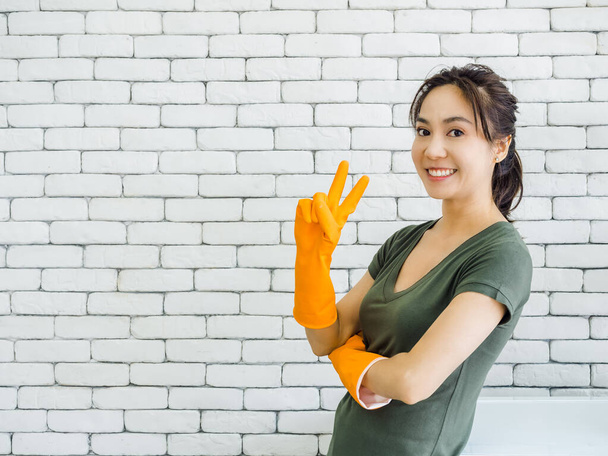 Ευτυχισμένο χαρούμενο χαμόγελο ασιατική γυναίκα, νοικοκυρά φορώντας πορτοκαλί γάντια καουτσούκ δείχνει δύο δάχτυλα, πινακίδα νίκη κοντά στο πλυντήριο σε λευκό φόντο τούβλο τοίχο στο δωμάτιο πλυντηρίων με αντίγραφο χώρο. - Φωτογραφία, εικόνα