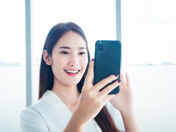 若い幸せなアジアの女性は会話の携帯電話を使用している間挨拶のために手を振っている。美しい女性は白いシャツを着てオンラインでビデオ通話を持っているスマートフォンを保持. - 写真・画像