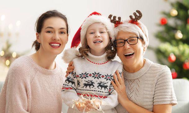 Merry Christmas and Happy Holidays! Cheerful kid, mom and granny. Parents and little child having fun near tree indoors. Loving family at home. - 写真・画像