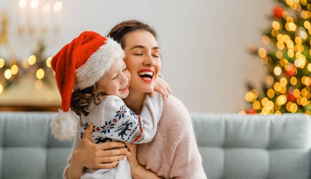 Merry Christmas and Happy Holidays! Cheerful mom and her cute daughter girl embracing. Parent and little child having fun near tree indoors. Loving family at home.  - Foto, immagini