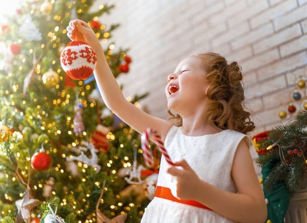 Merry Christmas and Happy Holidays! Cute little child girl is decorating the tree indoors. The morning before Xmas.  - Фото, зображення