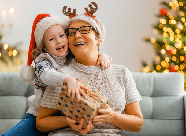 Merry Christmas and Happy Holidays! Cheerful grandma and her cute grand daughter girl exchanging gifts. Granny and little child having fun near tree indoors. Loving family with presents in room. - Foto, imagen