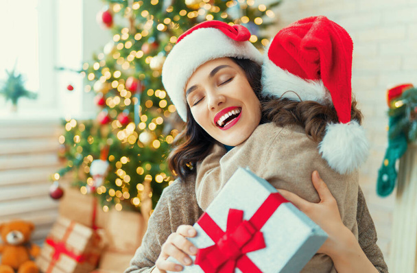 Merry Christmas and Happy Holidays! Cheerful mom and her cute daughter girl exchanging gifts. Parent and little child having fun near tree indoors. Loving family with presents in room.    - Foto, imagen