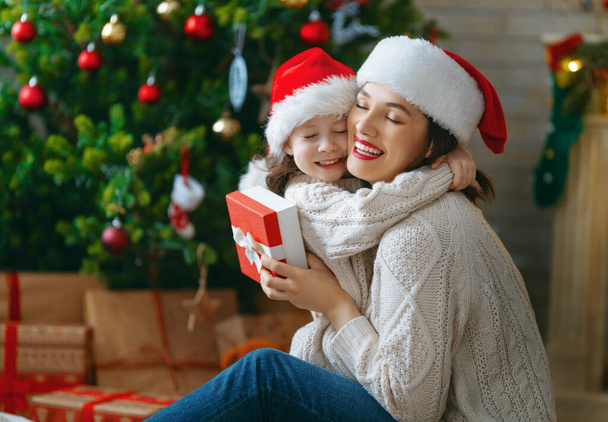 Merry Christmas and Happy Holidays! Cheerful mom and her cute daughter girl exchanging gifts. Parent and little child having fun near tree indoors. Loving family with presents in room.    - Foto, Imagen