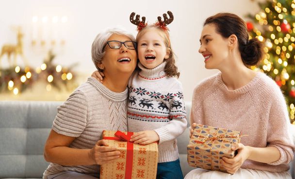 Merry Christmas and Happy Holidays! Cheerful kid presenting gifts to mom and granny. Parents and little child having fun near tree indoors. Loving family with presents in room. - 写真・画像