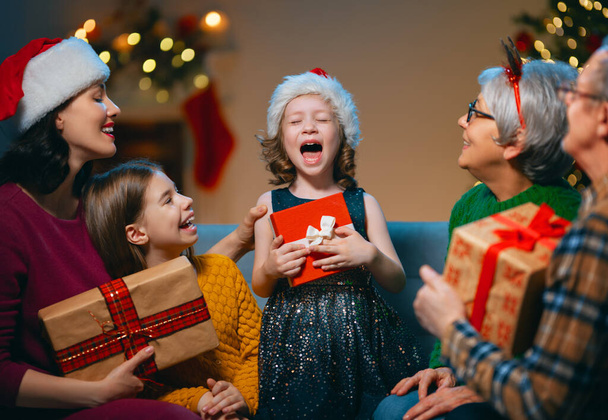 Merry Christmas and Happy Holidays! Grandma, grandpa, mum and children exchanging gifts. Parents and daughters having fun near tree indoors. Loving family with presents in room. - 写真・画像