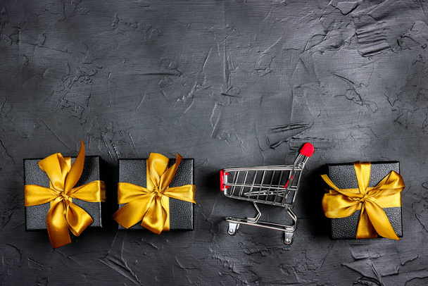 Shopping trolley and gift boxes with gold ribbons on black background. Holiday shopping and sale or black friday concept. Top view with copy space. - Foto, Bild
