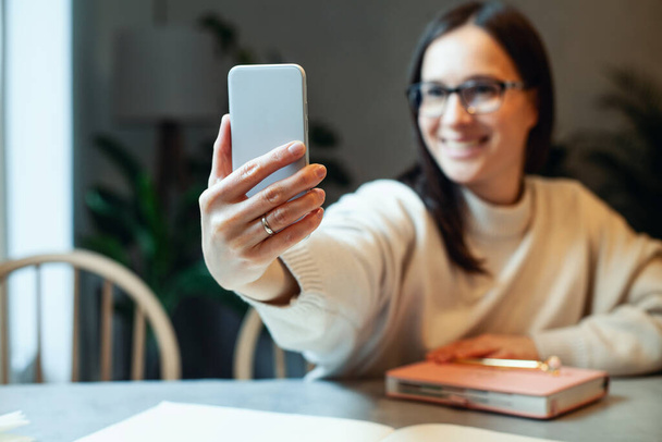 Happy young woman in eyeglasses, warm sweater is taking selfie photo on smartphone while sitting and drinking coffee at cafe with pink diary book and pen. Technology. Blogging and leisure concept. - Photo, Image