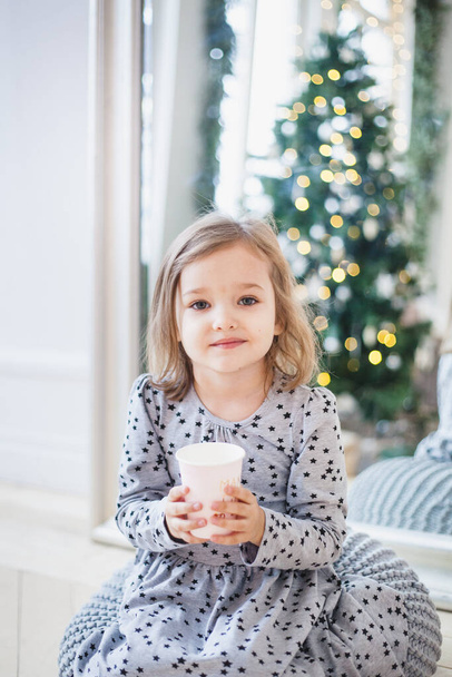 The room is decorated for the new year, the girl is sitting near the mirror, holding a glass or mug with a drink, milk or juice, Christmas - Foto, Imagem