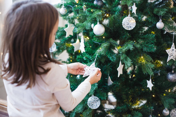 The room is decorated for the new year, the girl decorates the Christmas tree, balls, nests, waiting for Christmas, white color, baby's hands - Фото, изображение