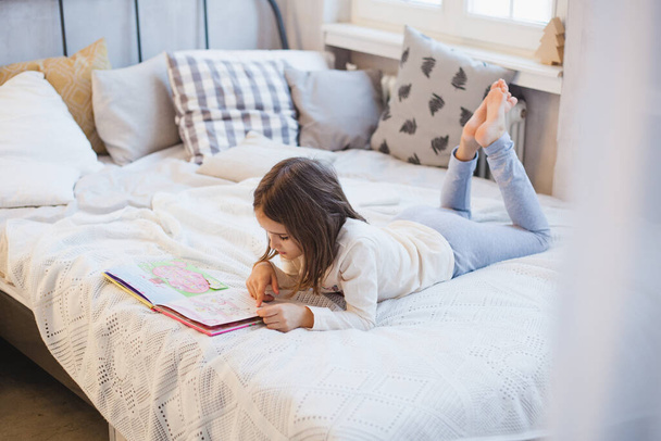 The room is decorated for the new year, the baby girl is lying on the bed and reading a book, learning, fairy tales, fun and childhood - Foto, Imagem
