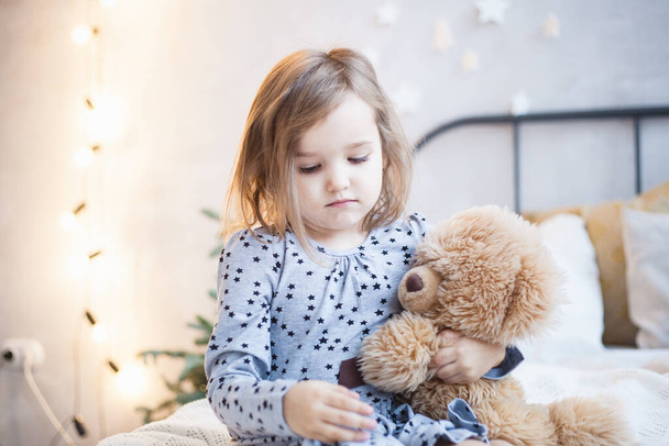 The room is decorated for the new year, a girl is sitting on the bed with a bear, a plush toy is playing school, reading a book, leafing through, a preschooler - Photo, Image
