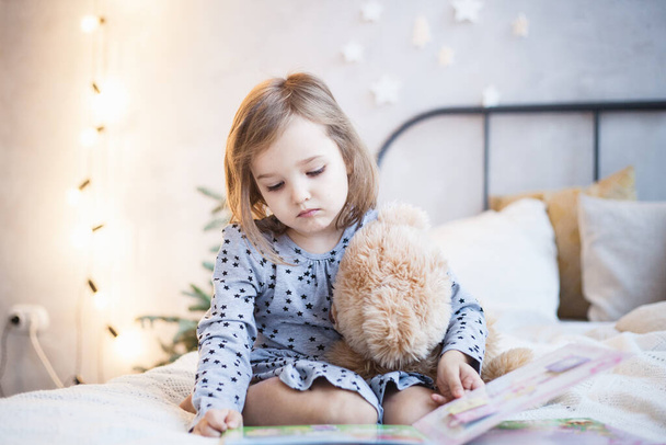 The room is decorated for the new year, a girl is sitting on the bed with a bear, a plush toy is playing school, reading a book, leafing through, a preschooler - Foto, immagini