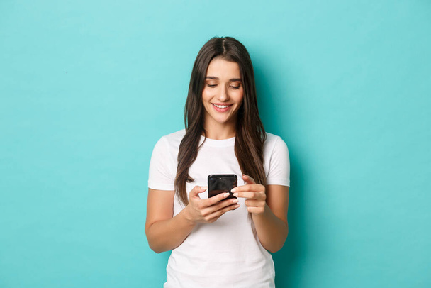Fiatal, modern nő fehér pólóban, mobiltelefonnal. Lány üzenetküldés és mosolygós, álló felett kék háttér - Fotó, kép