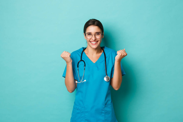 Coronavirus, pandemic and social distancing concept. Image of beautiful female doctor, triumphing and celebrating victory, saying yes and smiling, achieve goal over blue background - Photo, Image