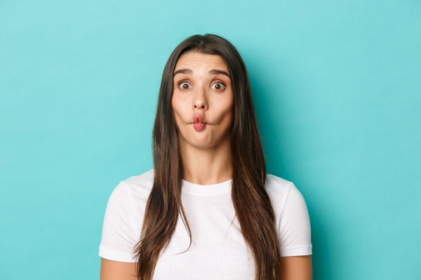 Close-up of silly brunette woman, sucking lips and showing funny faces, standing over blue background - Photo, Image