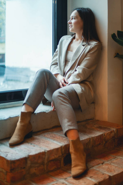 Fashionable young woman with long dark hair sits by the window. - Фото, зображення