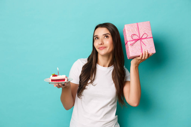 Image of attractive birthday girl, holding b-day cake and shaking gift box, guessing what inside, standing over blue background - Foto, afbeelding
