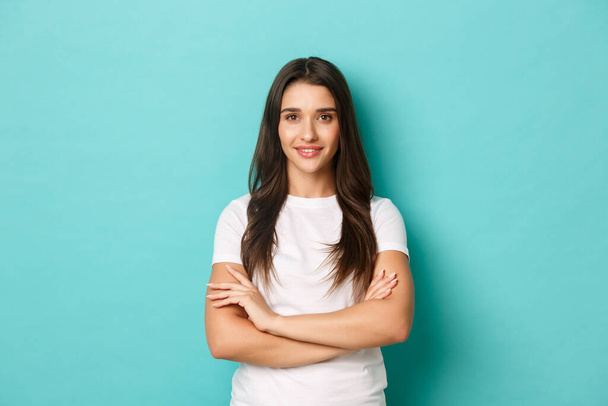 Confident young woman in white t-shirt standing with crossed arms on chest, looking at camera self-assured, standing over blue background - Photo, image