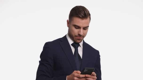 sexy young man wearing navy blue suit, holding phone, texting and smiling, making ok gesture, standing isolated on white background in studio - Footage, Video