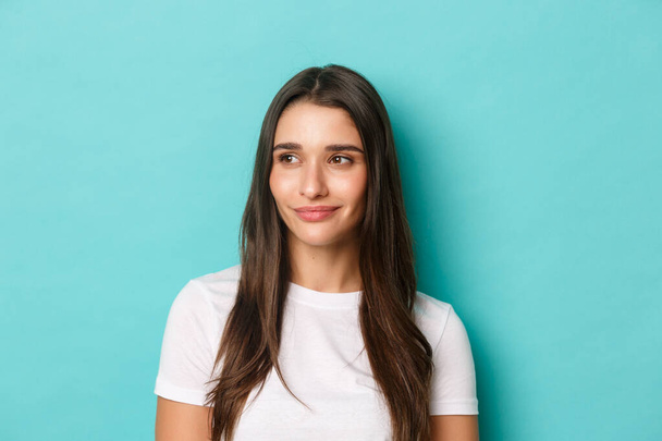 Close-up of attractive modern woman with long dark hair, wearing white t-shirt, looking left and smiling, standing over blue background - Photo, Image