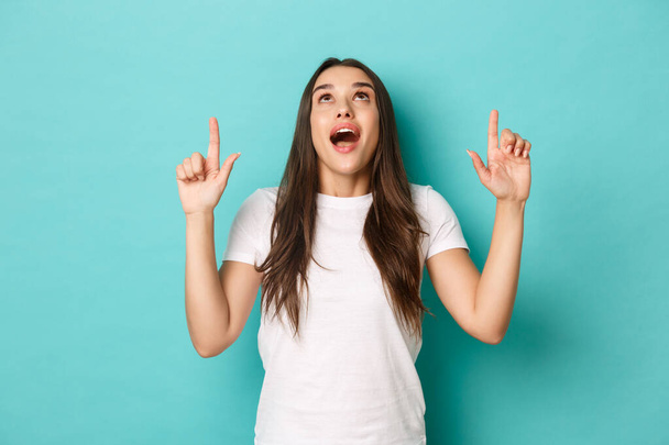 Portrait of amazed pretty woman in white t-shirt, looking and pointing fingers up with fascinated expression, standing over blue background - Foto, Imagen